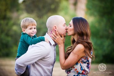 A couple sharing a kiss while their young son rides piggyback outdoors.