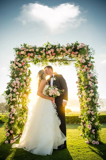Bride and groom kissing under a floral arch at sunset.