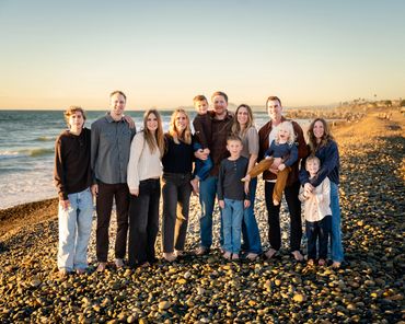 Large family posing barefoot on a rocky beach at sunset with smiles and casual attire.