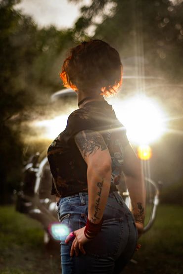 Person with tattoos and red hair backlit by bright light, standing outdoors near a motorcycle.