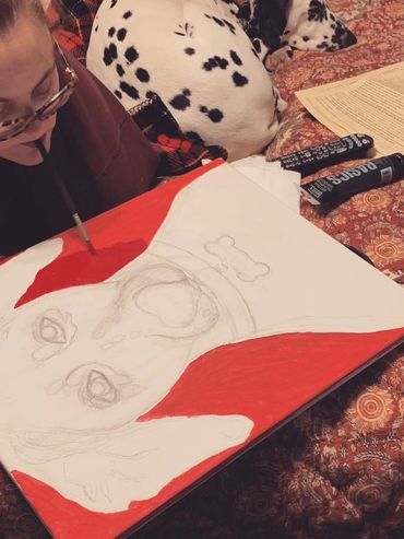 Summer Braddock painting red background on dog portrait by mouth. Her dalmatian cuddles nearby.