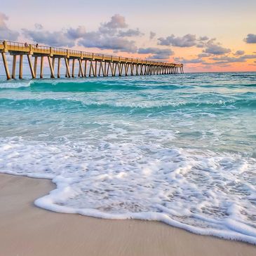 A sandy beach with gentle waves and a long pier at sunset.