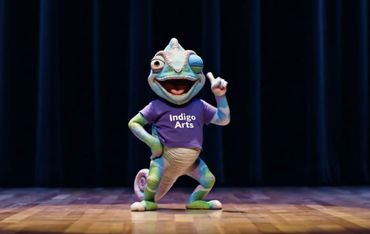 A colorful chameleon mascot wearing an "Indigo Arts" shirt poses on stage.