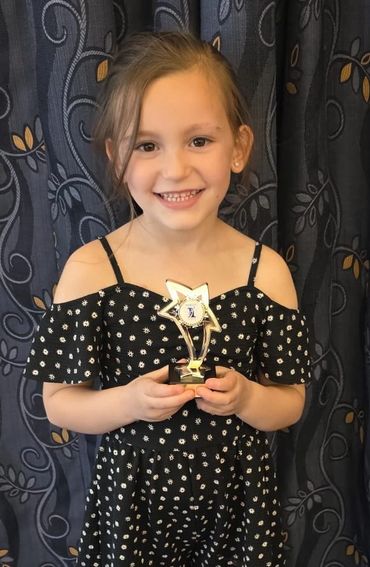 Smiling girl holding a star-shaped trophy, wearing a black dress with white flower patterns.