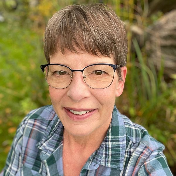Close-up of a smiling woman with short brown hair and glasses, wearing a green and blue plaid shirt