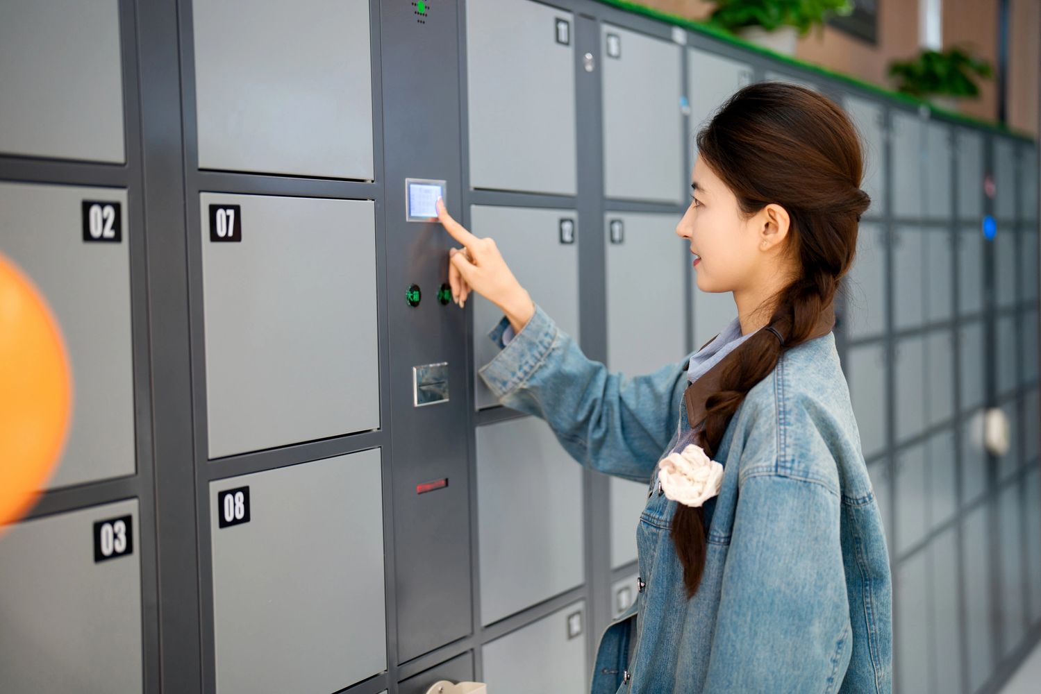 Woman with braided hair using a touchscreen on a row of numbered gray lockers indoors.