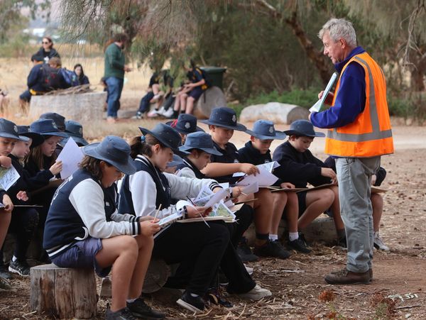 Students in hats sit outdoors, writing, as a man in an orange vest instructs.