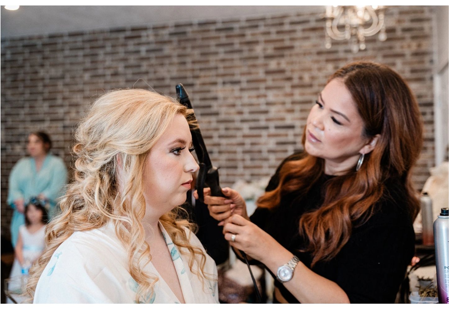 A hairstylist curling a woman's blonde hair indoors with a brick wall background.