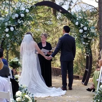 Bride and groom holding hands during outdoor wedding ceremony under floral arch.