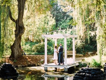 A couple stands under a white gazebo surrounded by lush greenery and water.