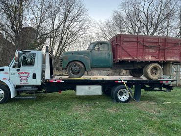 A vintage green truck with a red cargo bed loaded on a white flatbed tow truck.