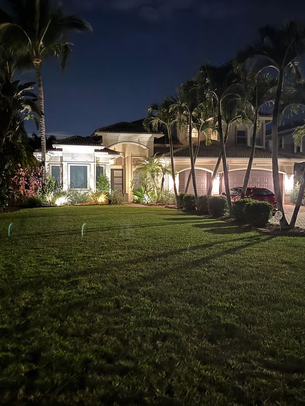 A well-lit house with palm trees and a lush green lawn at night.