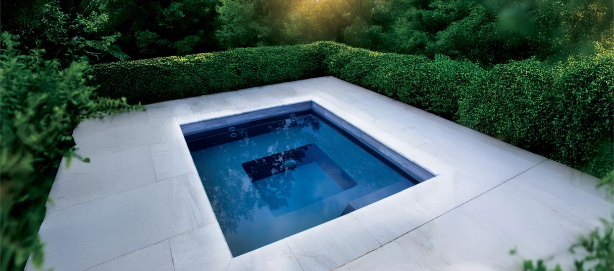 A serene, modern square pool surrounded by lush green hedges and white stone tiles.