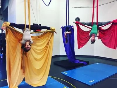 Three people practicing aerial silks upside down with colorful fabrics indoors.