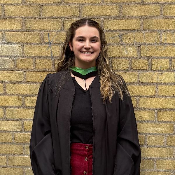 Young woman smiling in graduation gown against a brick wall.