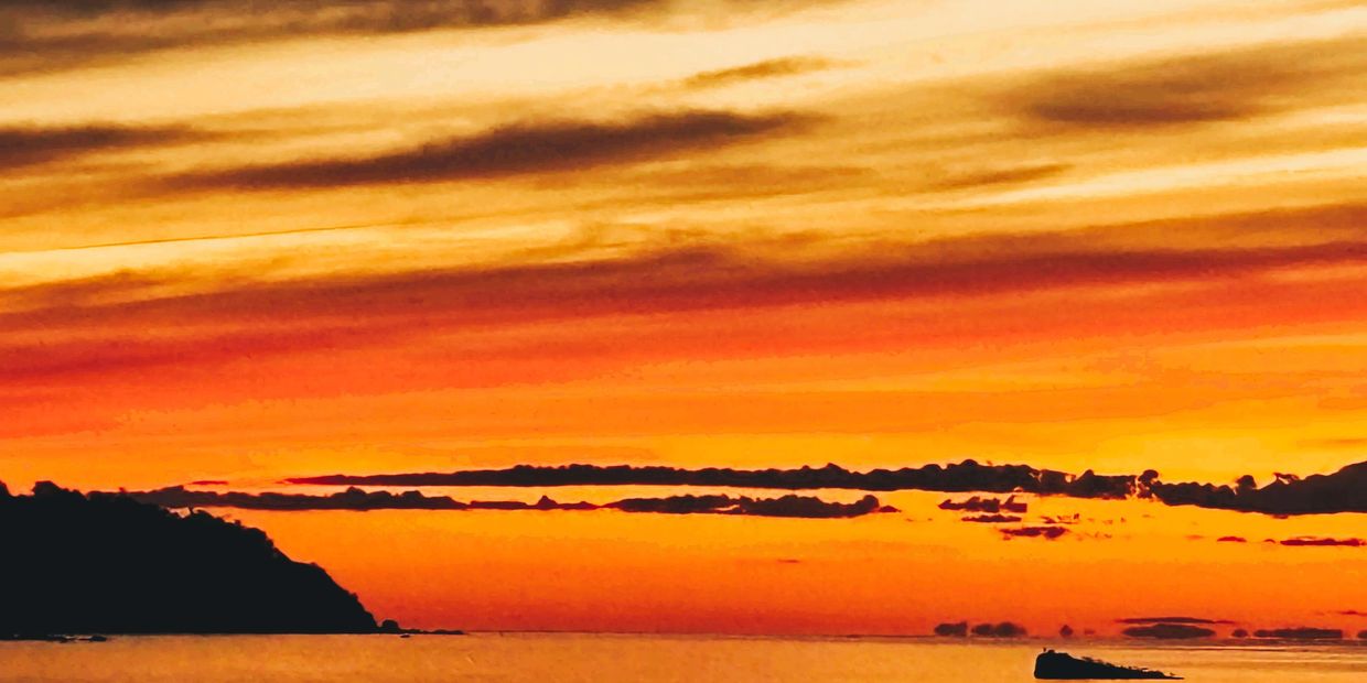 Vibrant orange sunset over calm sea with silhouetted land and scattered clouds.