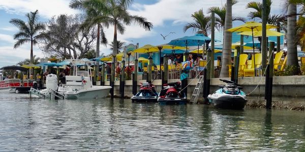 Boats and jet skis docked near a lively waterfront with colorful umbrellas and palm trees.