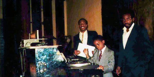 Three men in suits around a DJ turntable.