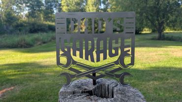 Metal sign reading "Dad's Garage" with crossed wrenches, outdoors on a tree stump.