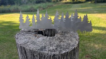 Metal silhouette of pine trees placed on a weathered wooden stump outdoors.