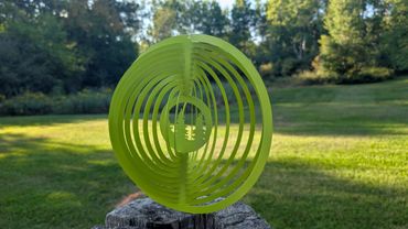 A green circular metal wind spinner with tree cutouts on a wooden post.