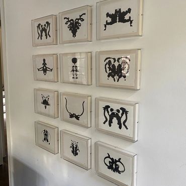 Twelve framed black inkblot art pieces arranged neatly on a white wall.