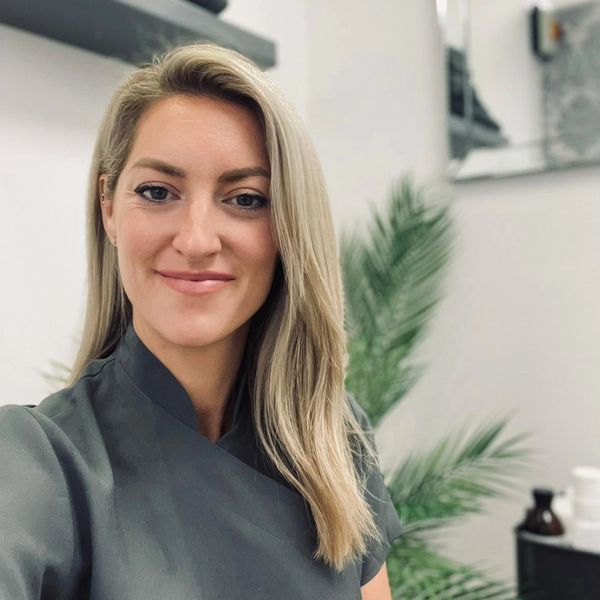 Smiling woman with blonde hair in a gray top, indoors with green plants in the background.