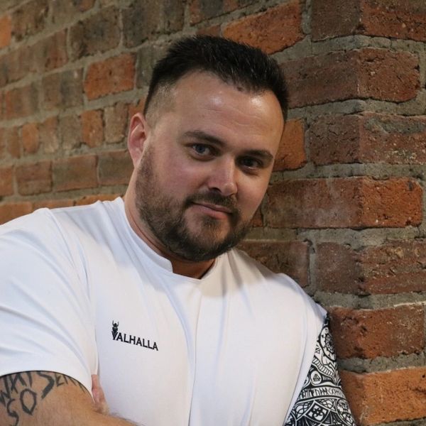 Man with tattoos wearing a white Valhalla t-shirt leaning against a brick wall.