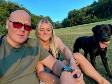 A man and woman sit on grass with a black dog holding a ball.