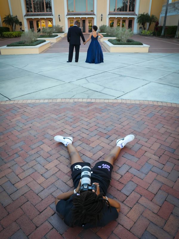 Photographer lying on brick pavement capturing a couple holding hands in front of a building.
