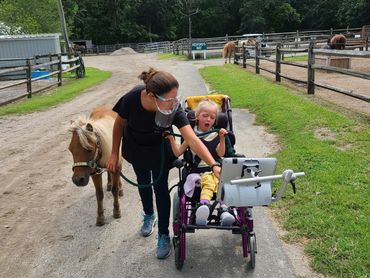 Child in a wheelchair using an eye gaze AAC system while working with a speech therapist and horse.