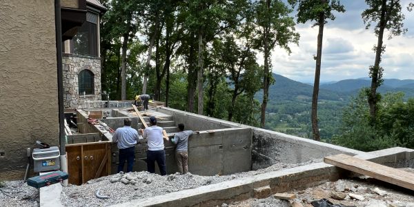Three workers inspecting a concrete foundation near a house in a wooded mountainous area.
