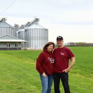 Couple standing in a green field near silos, smiling.