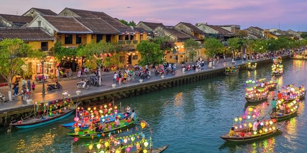 Colorful lantern-lit boats float on a river beside lively streets at dusk.