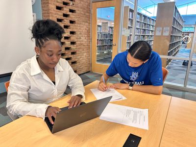 Two students studying at a library table with laptop and papers.