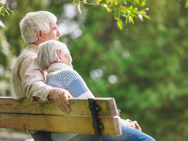 An older couple, leaning against each other on a park bench. 