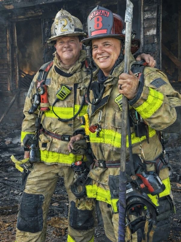Two firefighters in gear standing in front of a burnt building, smiling.