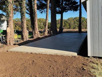 Newly poured concrete slab next to a grey shed and trees.