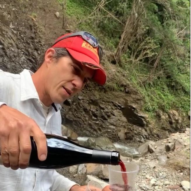 Man pouring red wine into a plastic cup outdoors by a rocky stream.