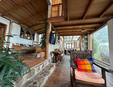 Cozy rustic interior with wooden furniture, colorful cushions, and large windows overlooking greenery.