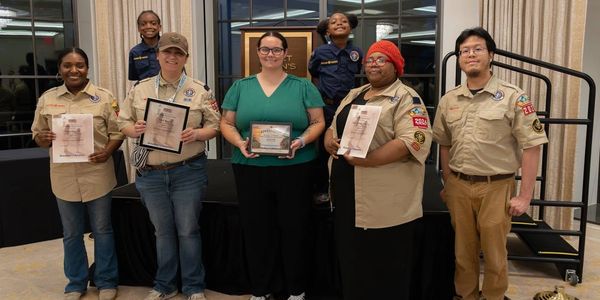 Five adults stand in a row and wear variations of the tan Scouts BSA uniform. Four hold various awar