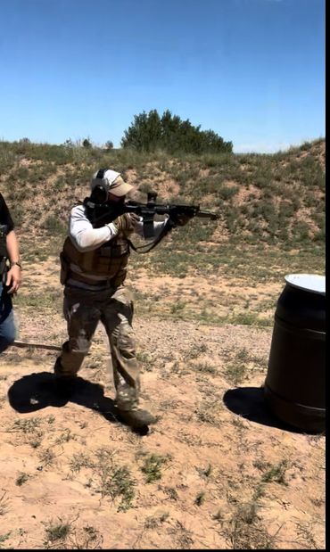 Person in tactical gear aiming rifle outdoors near a barrel.