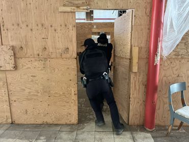Two law enforcement officers in tactical gear entering a wooden training structure.