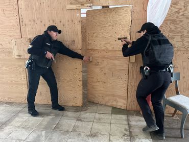 Two armed officers conduct a tactical training exercise behind plywood walls.