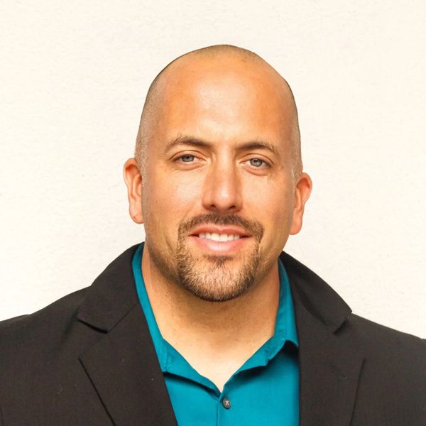 Smiling man with a shaved head in a black blazer and teal shirt.