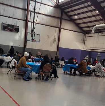 Community gathering in a gym with people seated at tables for a Coffee, Tea and Sanity event by Nurturing Possibilities