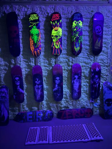 Glowing skateboards with neon skull and horror designs displayed on a textured wall under blue lighting.