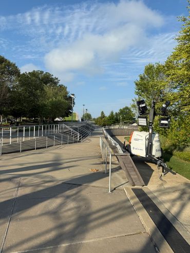 Outdoor pathway with railings, trees, and a mobile light tower under a blue sky.