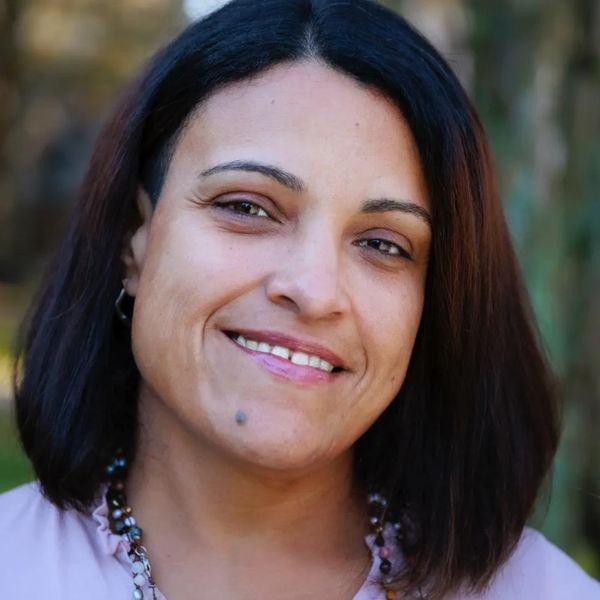 Smiling woman with dark hair and a beaded necklace outdoors.