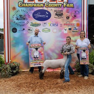 Three people holding banners with a sheep at the Champaign County Fair.